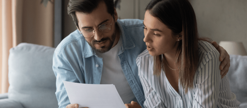A man and woman reviewing documents together. Man has his arm around the woman. Both are in a living room.