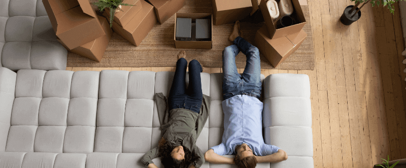 Overhead view of a couple lying on a couch after moving. Boxes and plants are nearby.
