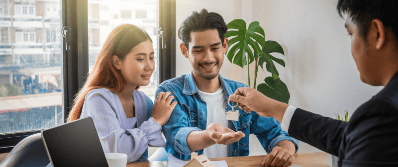 Man handing keys to a smiling couple in an office setting. Sunlight streams in.