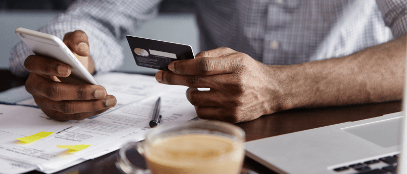 Person using phone and credit card, sitting at a table with documents, laptop, and coffee.