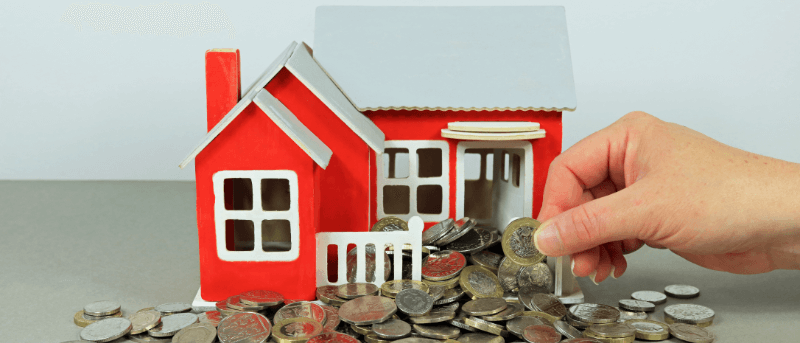 A red house model with a hand adding coins to a pile, symbolizing home savings.