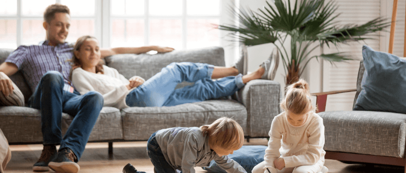 Family relaxing on a couch. Children playing on the floor, near a window.
