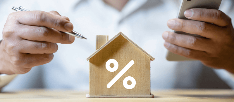 Person with pen and phone near a wooden house with a percentage symbol, symbolizing mortgage rates.
