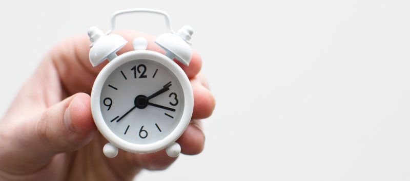 A hand holding a white alarm clock with the hands pointing to 2:15.