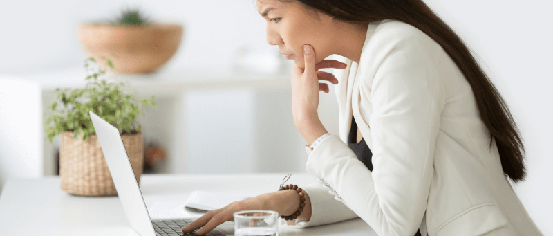 Woman in white blazer using laptop, hand on chin, in a bright office setting.