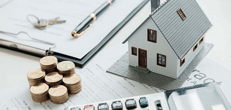 Miniature house next to paperwork, coins, keys, pen, and calculator, suggesting real estate investment or finances.