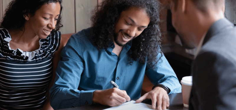 A man signs a document at a table with a woman watching as another man looks on.