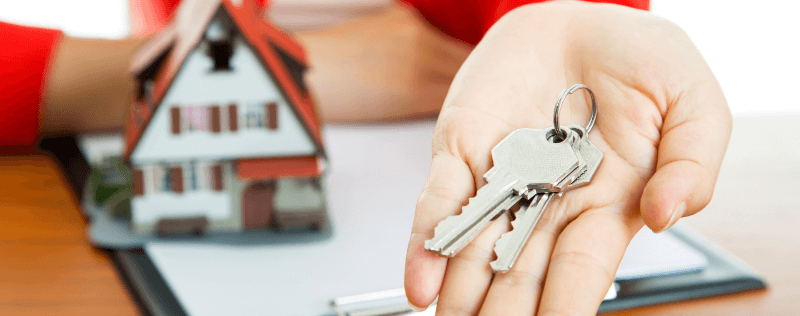 Person holding keys with a model house in the background, symbolizing home ownership.