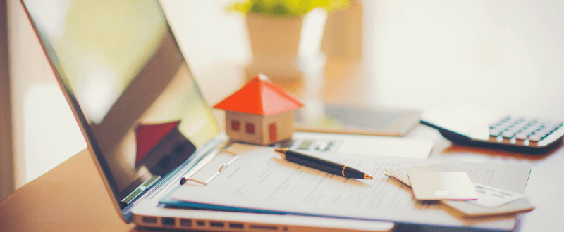 Laptop with a miniature house on top, paperwork, pen, and calculator, suggesting real estate or home finances.
