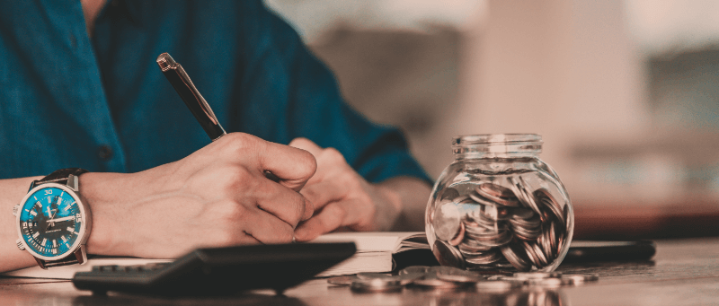 Person writing with pen, with jar of coins and calculator nearby, in an office setting.