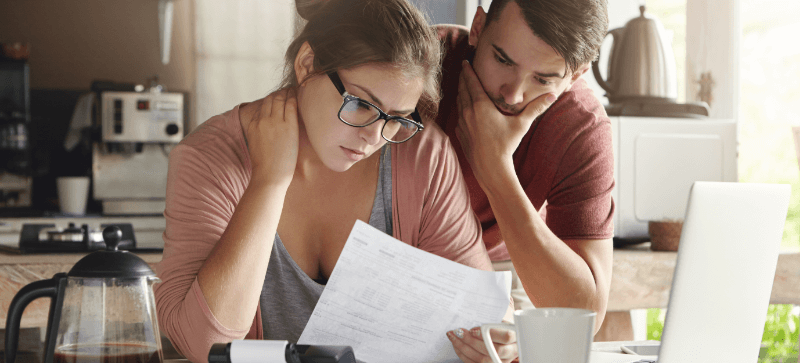 A concerned couple reviews paperwork at a kitchen table, likely focused on finances, with a laptop and coffee.