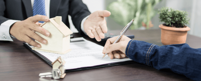 Person signing a real estate contract with a miniature house and keys on a table.