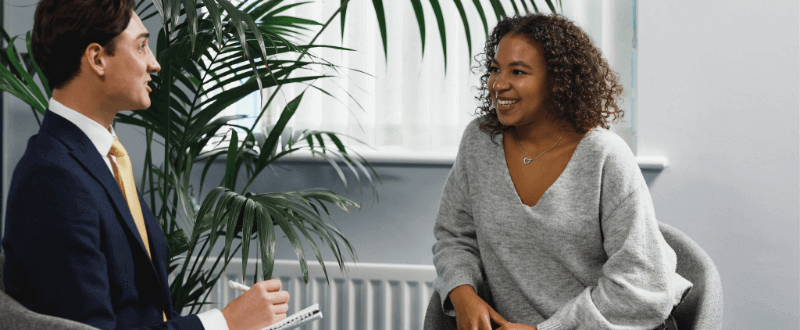A man in a suit interviews a woman in a gray sweater in a well-lit room.