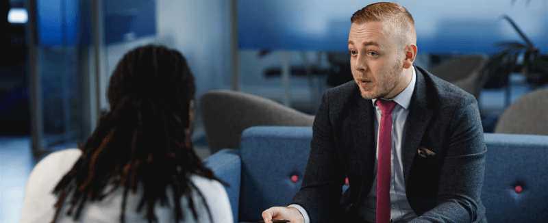 Man in suit talking to person with dreadlocks, seated in a modern office setting.