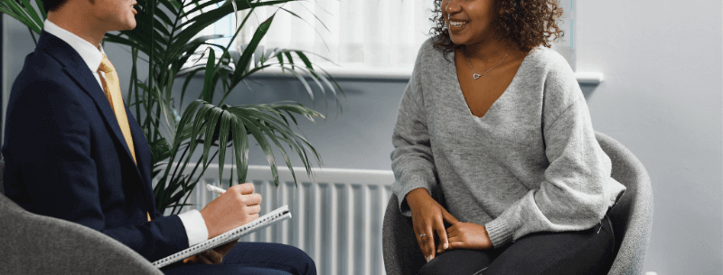 A therapist in a suit takes notes while a smiling woman talks. They sit in chairs in a room.