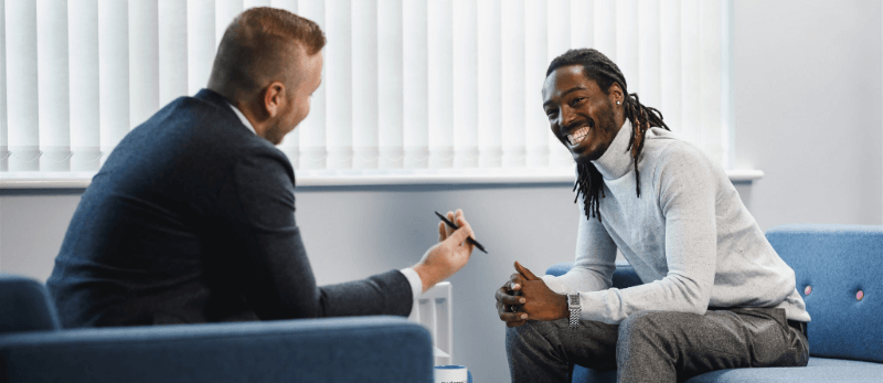Two men in an office setting, one in a suit holding a pen, smiling, and the other with dreadlocks, smiling.