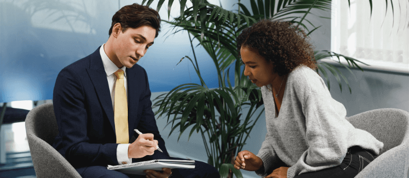 A man in a suit reviews paperwork with a woman in a sweater in an office setting.
