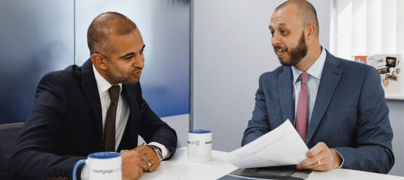 Two men in suits at a table. One holds papers, the other looks on. They are in an office setting.