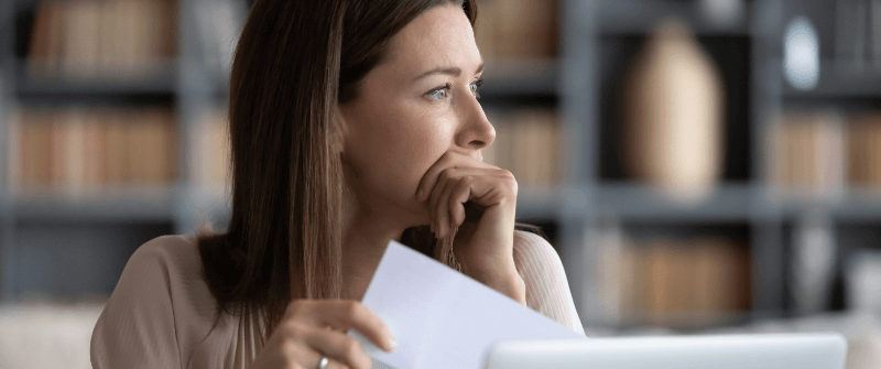 Woman looks thoughtful, holding paper, hand to her face, in an office setting with blurred background.