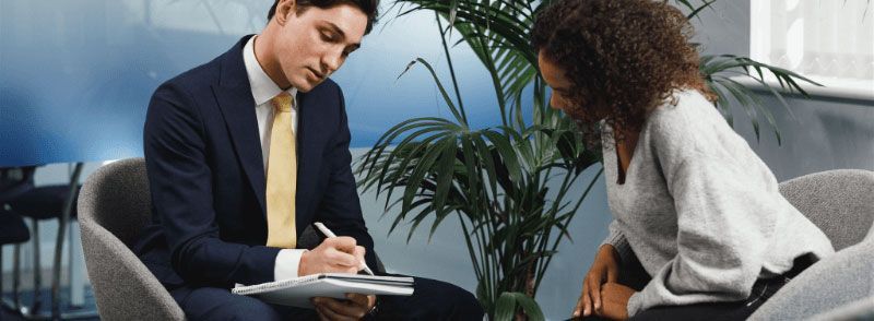 A man in a suit writes on a clipboard while consulting with a woman in a light-colored sweater.