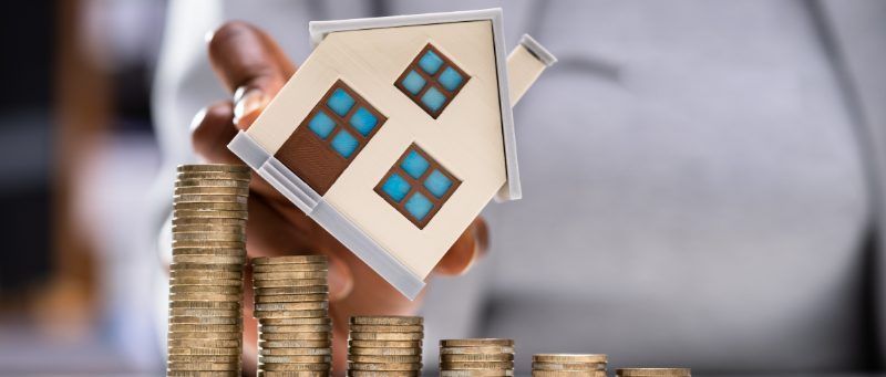 Person holding a model house over stacks of coins, illustrating property investment.