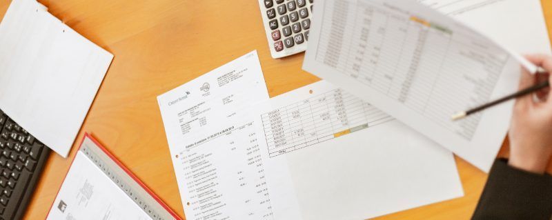 A person reviewing documents on a wooden desk with a calculator, papers, and a pen.