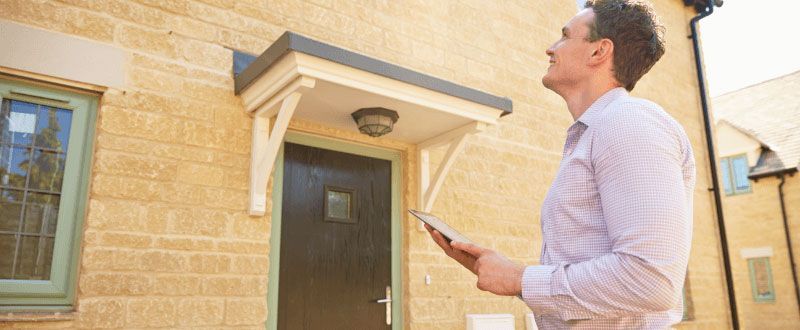 A man examines a house, holding a tablet. He wears a light shirt and is looking upwards.