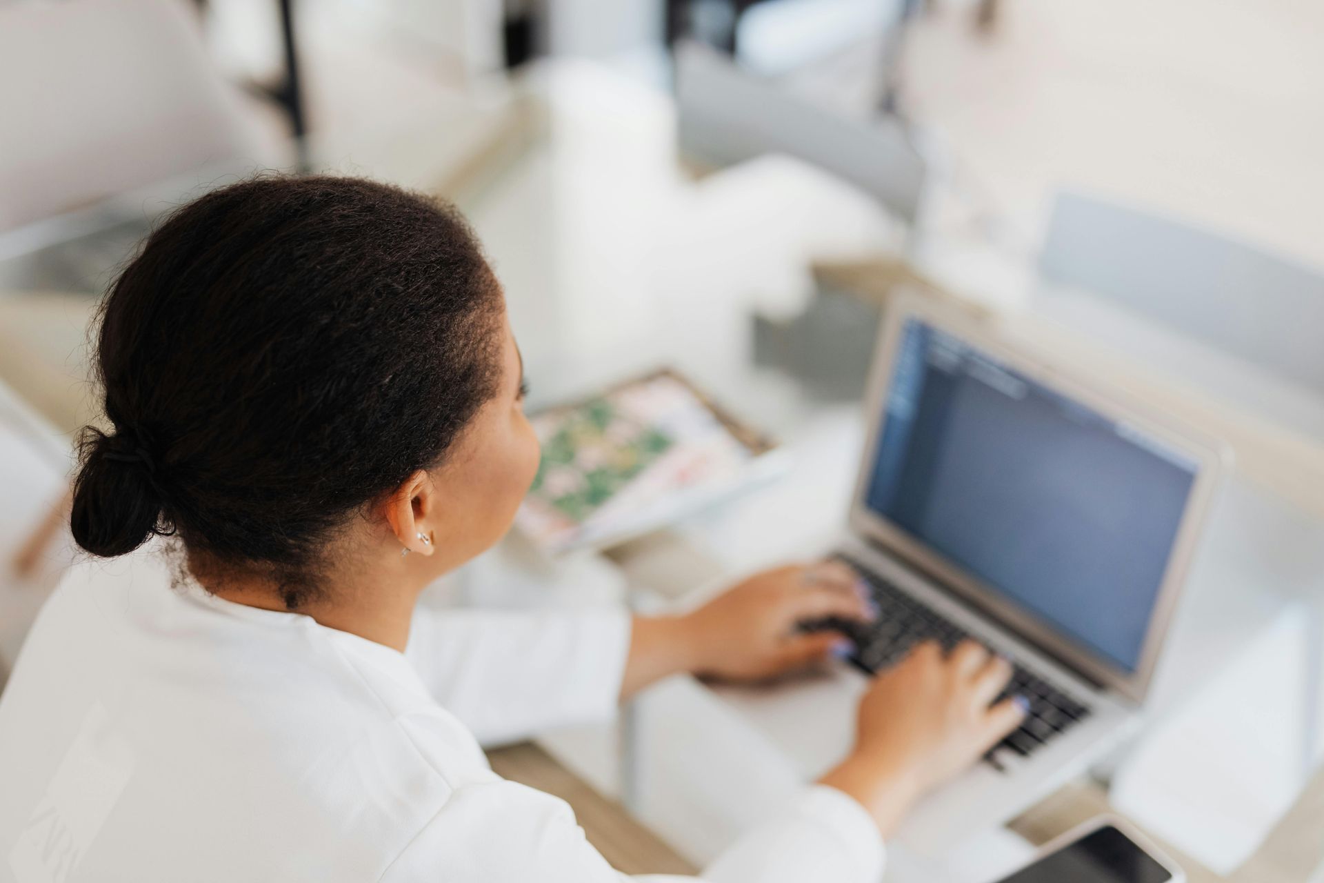 Woman with dark hair typing on a laptop at a table in a bright room.