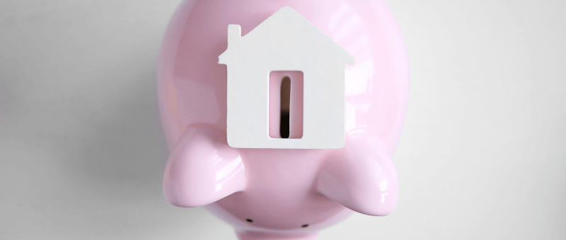 Pink piggy bank with a white house-shaped slot on top, symbolizing saving for a home.