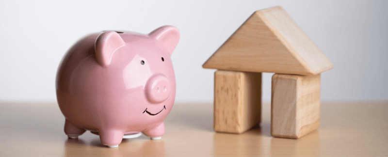 Pink piggy bank next to a wooden house made of blocks, symbolizing savings for a home.