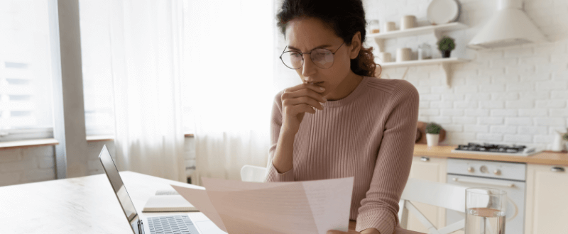 Woman wearing glasses thoughtfully reviewing papers in kitchen with laptop.