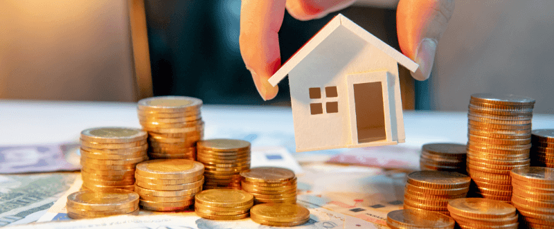 Hand holding a model house over stacks of coins and money, suggesting real estate investment.