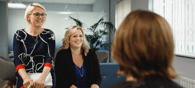 Three women in an office setting, smiling. Two are facing forward, one's back is to the camera.