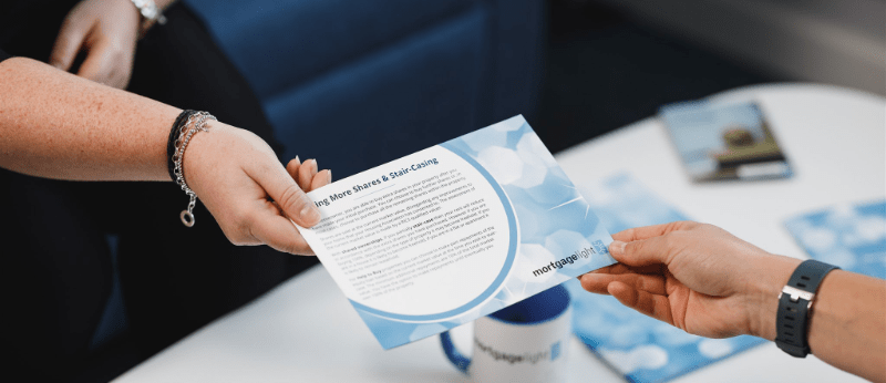 Two hands exchanging a blue brochure with text. A mug and other brochures are visible.