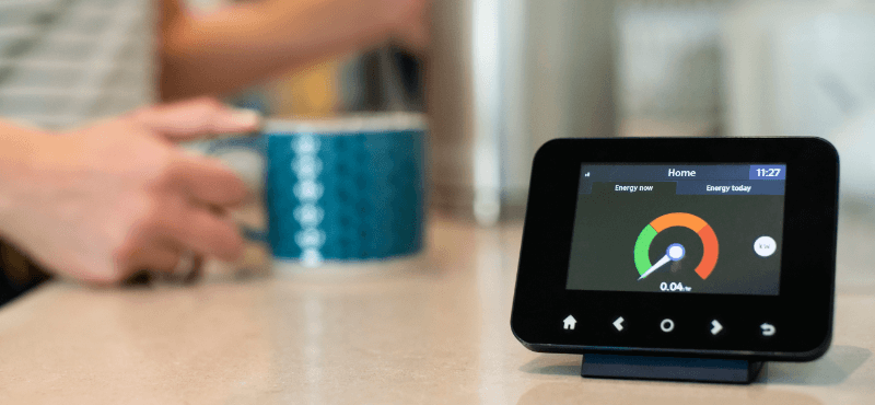 A digital energy monitor on a countertop, with a person holding a blue mug in the background.
