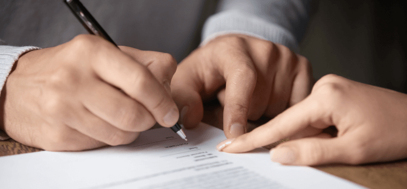 Person signing a document as another person points to a line on the paper.