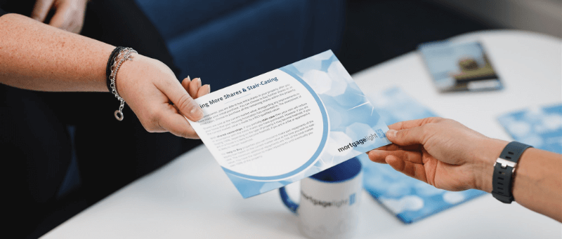 Hands passing a blue and white pamphlet with text. Two people are in an office setting.