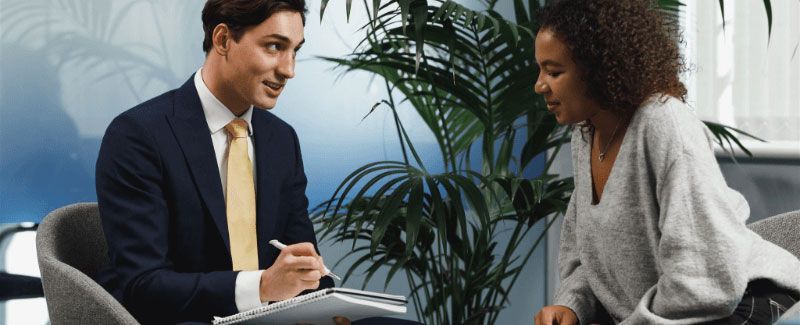 A man in a suit and a woman with curly hair sit and talk in an office setting, with a plant between them.