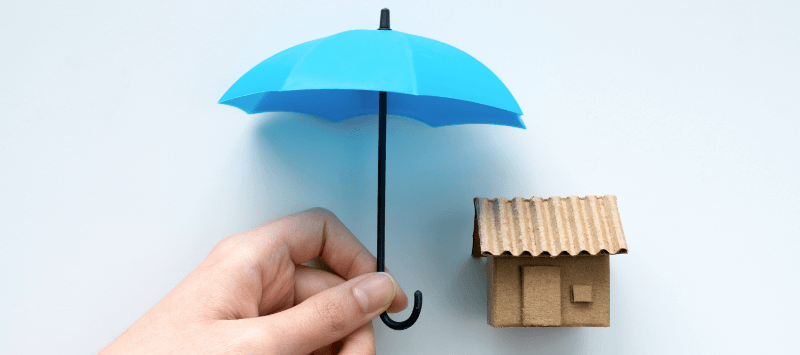 A hand holding a blue umbrella protects a miniature cardboard house.
