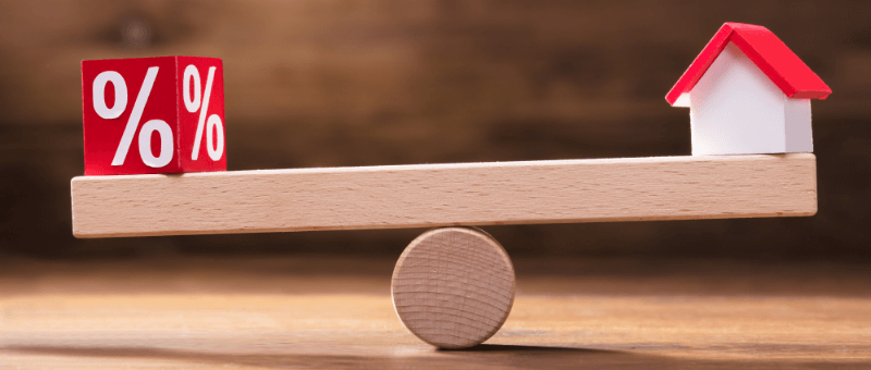 A seesaw balances a percentage cube and a miniature house, representing interest rates and housing.
