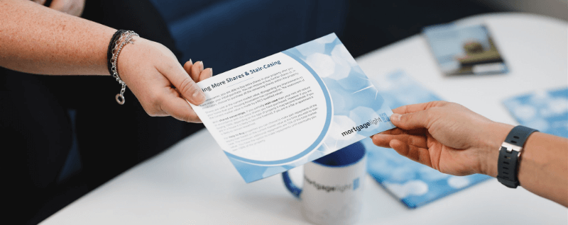 Two hands passing a blue flyer with text, over a white table. A mug is visible.