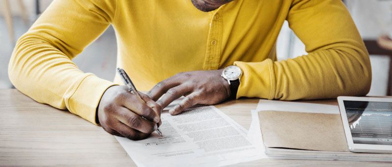 A person writing on paper, wearing a yellow shirt and watch, on a wooden table.