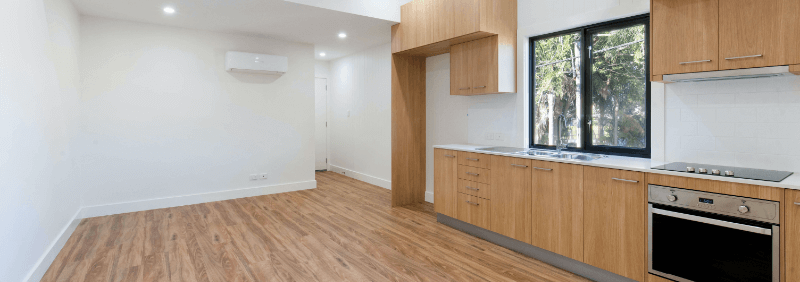 Empty kitchen with wooden cabinets, white countertops, oven, and window overlooking greenery.