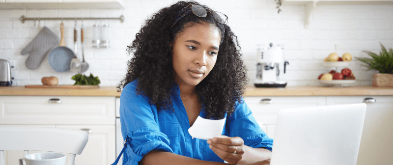 Woman in blue shirt examining a receipt while using a laptop in a kitchen.