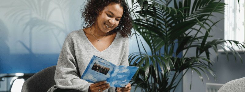 Woman in a gray sweater smiles while reading a brochure indoors with a plant in the background.