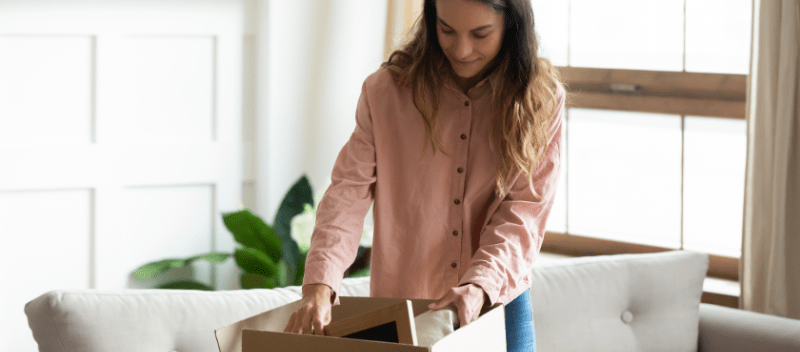 Woman unpacking a cardboard box in a living room. She wears a pink shirt and smiles, surrounded by natural light.