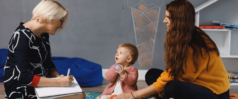 A baby with a pink cardigan sits between two women; one writes, the other smiles and touches the baby's hand.