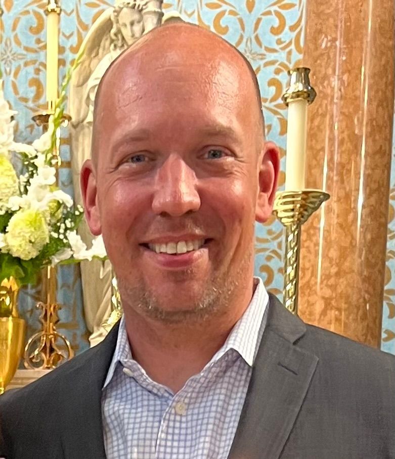 A bald man in a suit is smiling in front of candles and flowers.
