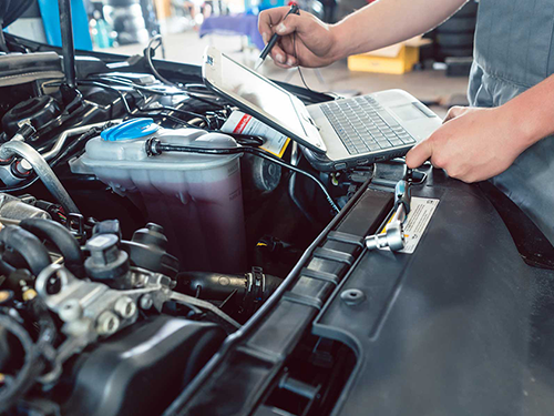 A man is working on the engine of a car while using a laptop.