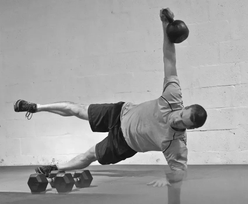 Homme en planche latérale avec kettlebell à un bras, des haltères posés à proximité sur le sol.